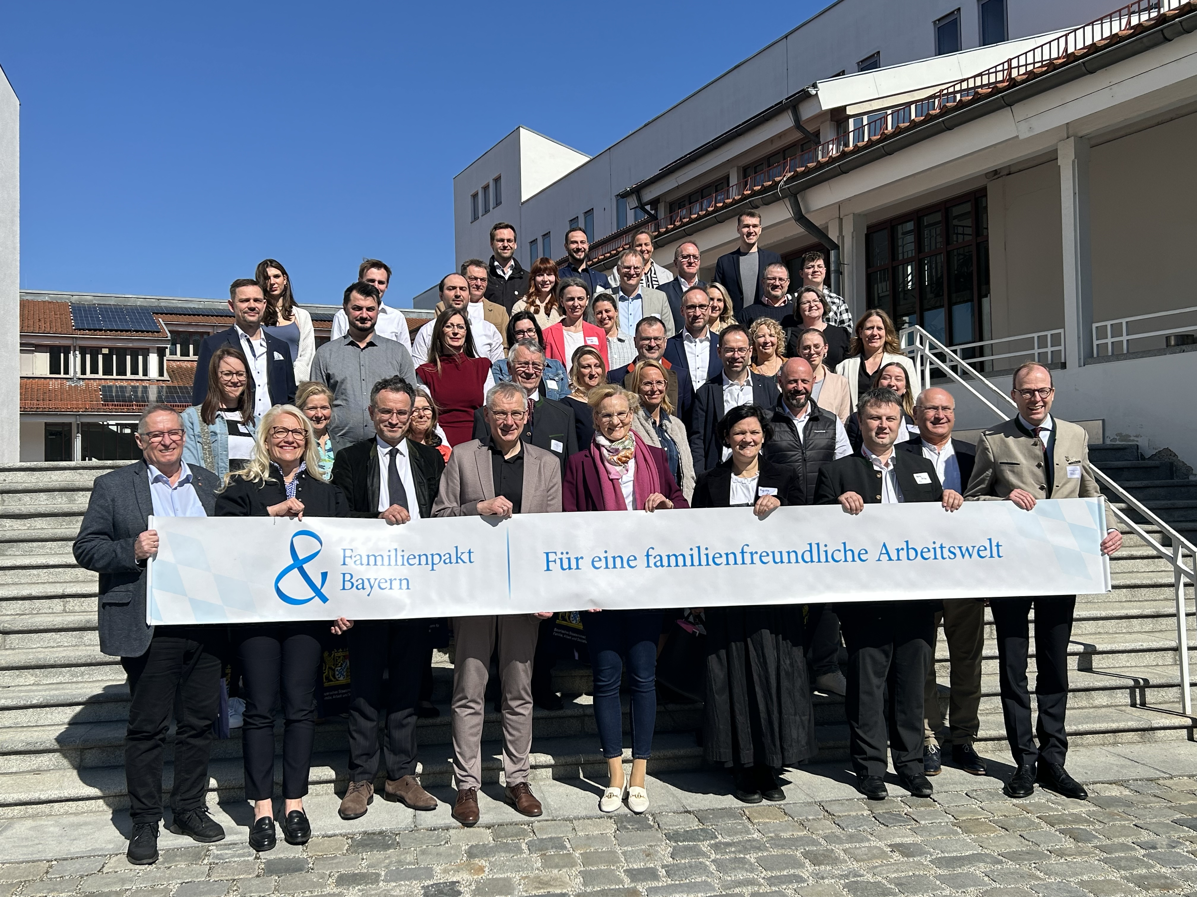 Eine Gruppe neuer Mitglieder des Familienpakts stehen zusammen. Sie präsentieren ein Banner mit der Aufschrift "Familienpakt Bayern".